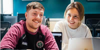 Two students, a male and female , itting in a computer lab working on a laptop and smiling.