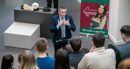 Mr Ciaran McLaughlin, Ulster GAA President talking to a group of students at the Ulster GAA student bursary awards.  Ciaran talking at a podium with hands pointed to his chest and we can see back of the students sitting and listening to his speech.