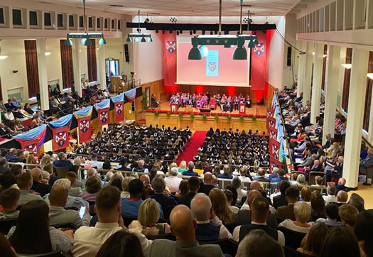 Graduation ceremony at Whitla Hall, Queen's University.  Taken from the balcony with family and friends of the students looking on and the graduates of the day on the lower floor space, seated with their graduation gowns on.  On the stage in front of them are the faculty of Queens and some of St Mary's taking part in the graduation ceremony.