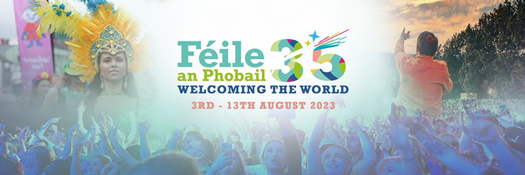 Féile an Phobail 35, 3rd - 13th August, Welcoming the World banner.  The bottom of the banner shows a crowd of people smiling with their hands in the air, from one of the concert nights. On the right hand of the banner shows the back of a person with their left hand in the air and singing to a crowd and the right hand of the banner shows a female with an elaborate teal head dress with yellow feathers from one of the previous years carnival parade.